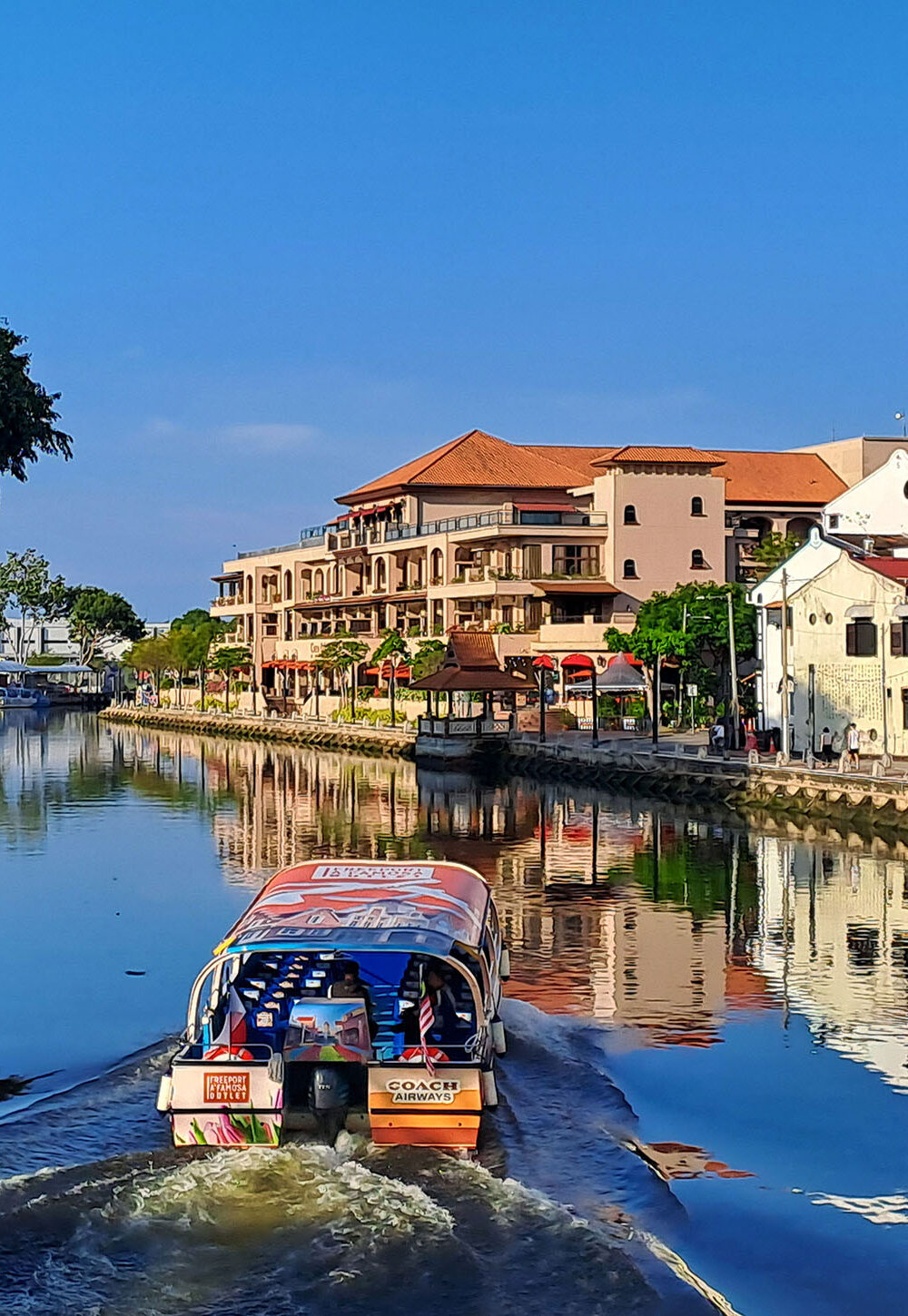 A sunny morning scene in Melaka of a boat on the river lined with buildings and trees