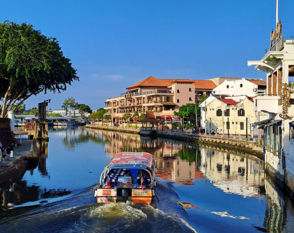 A sunny morning scene in Melaka of a boat on the river lined with buildings and trees