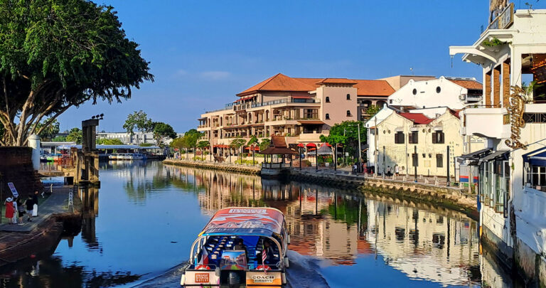A sunny morning scene in Melaka of a boat on the river lined with buildings and trees
