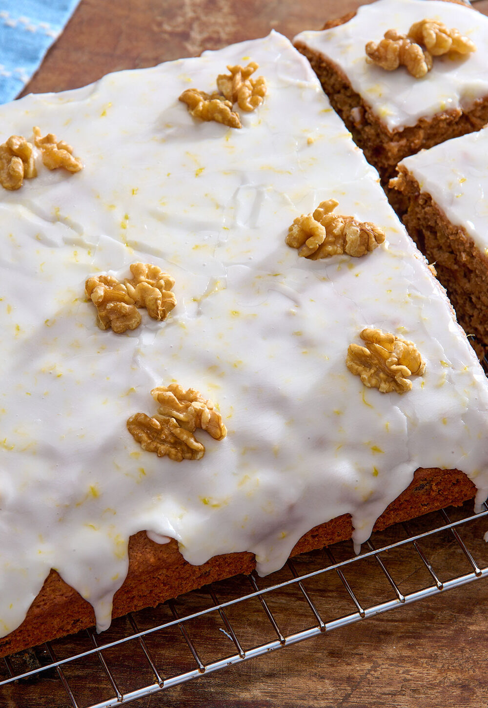 Angled shot of an iced date and walnut traybake, topped with whole walnut pieces, with three pieces cut out. The cake is sat on a cooling rack on top of a wooden table.