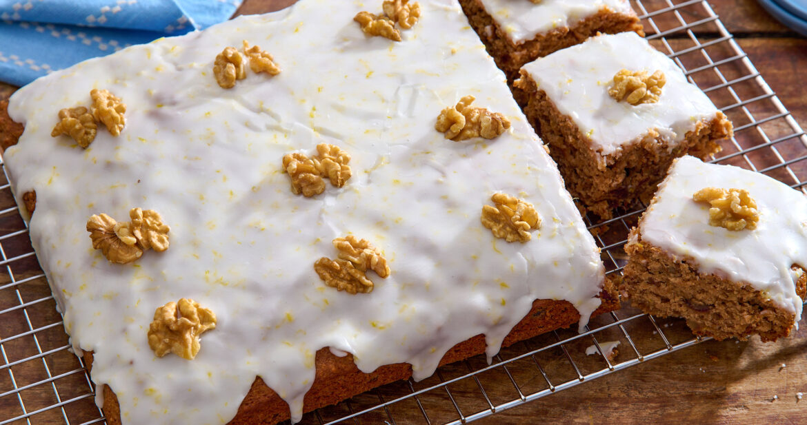 Angled shot of an iced date and walnut traybake, topped with whole walnut pieces, with three pieces cut out. The cake is sat on a cooling rack on top of a wooden table.