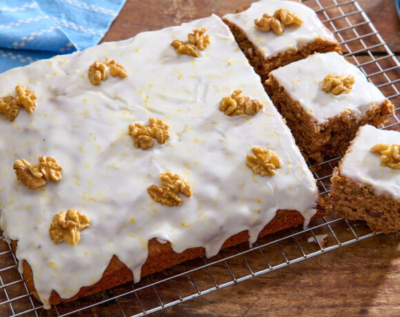 Angled shot of an iced date and walnut traybake, topped with whole walnut pieces, with three pieces cut out. The cake is sat on a cooling rack on top of a wooden table.