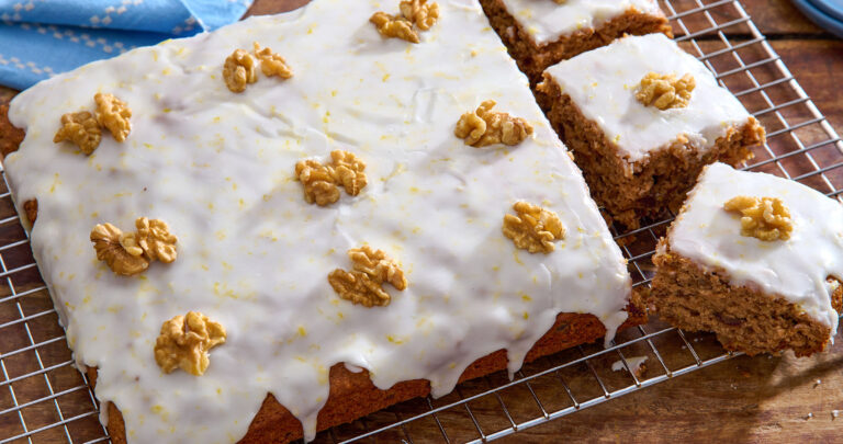 Angled shot of an iced date and walnut traybake, topped with whole walnut pieces, with three pieces cut out. The cake is sat on a cooling rack on top of a wooden table.